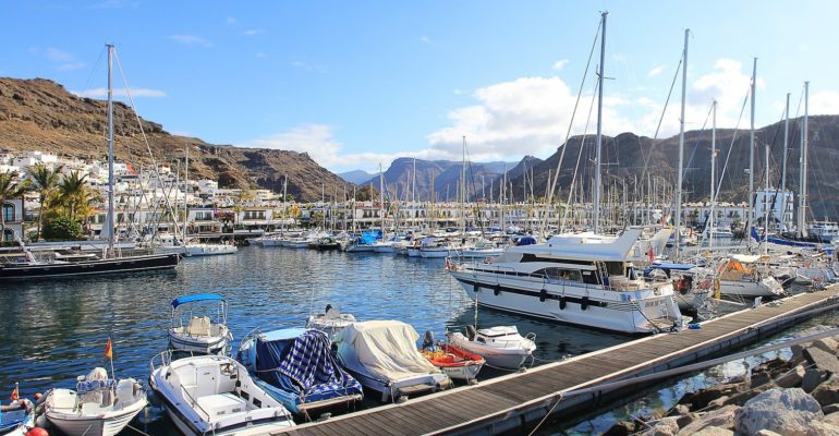 Many boats docked in a waterfront harbor with stunning mountain views, perfect for those looking to buy a property in Gran Canaria.