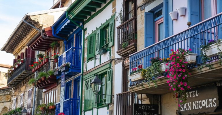 Set up your bed and breakfast in Spain and experience the vibrant life of a B&B manager surrounded by a colorful row of buildings adorned with balconies and flower pots.