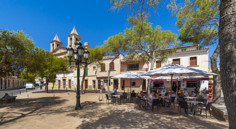 A town square with tables and chairs in front of a church, near a new build apartment in Mallorca.