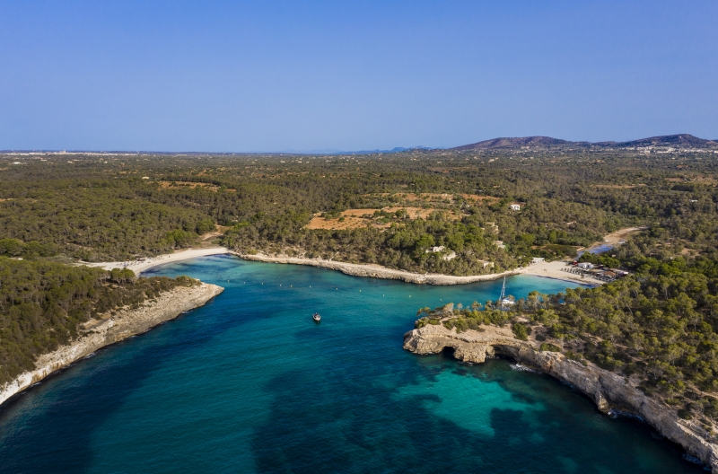 An aerial view of a beach near a new build apartment in Cala d'Or with blue water and trees.