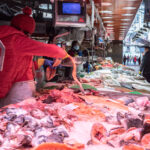 A vendor in a red jacket arranges fish and seafood on a long display counter at an indoor market along the Costa de Barcelona, with other people and stalls visible in the background.