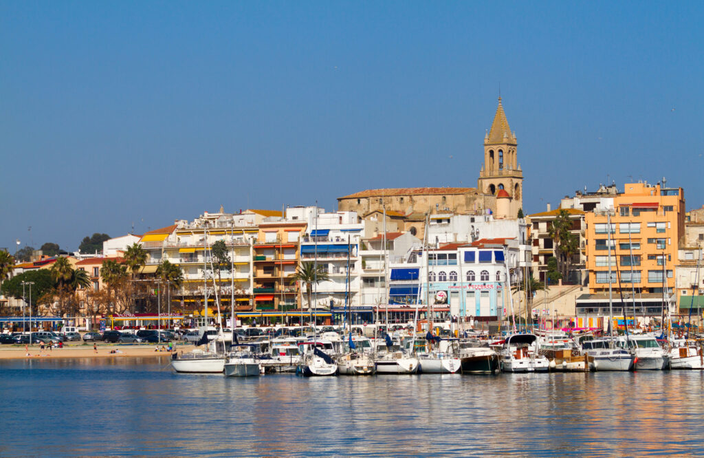 A waterfront view of colorful buildings and a church tower behind a marina filled with docked boats under a clear blue sky in Costa Brava, Spain.