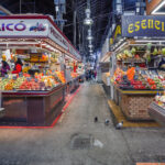 Colorful indoor market with fruit and vegetable stalls on either side, vendors arranging produce, and pigeons walking the aisle—a must-see spot featured on any Barcelona map for those exploring the best things to do on the Costa de Barcelona.
