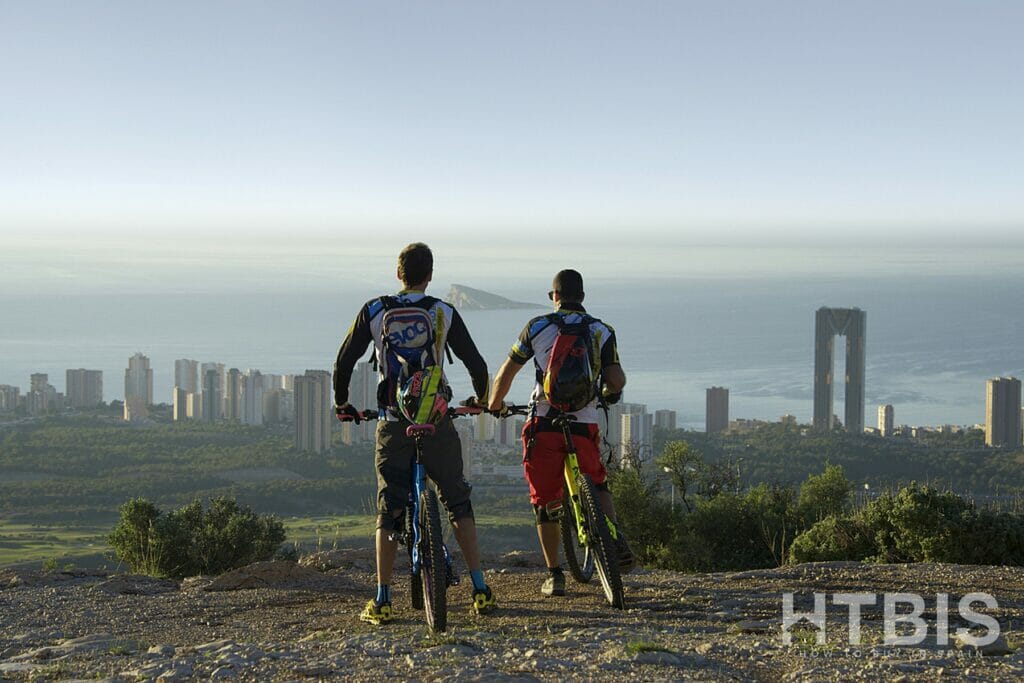 Two mountain bikers standing on top of a mountain overlooking Alicante, with views of the new build apartments and properties for sale.