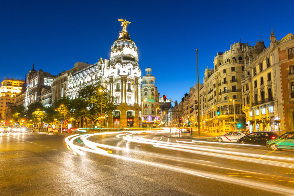 Nighttime view of a busy street with light trails from moving vehicles, showcasing an illuminated historic building with a dome and statue on top, surrounded by other ornate buildings. Discover the Spanish Costas and cities while soaking in the vibrant urban nightlife.