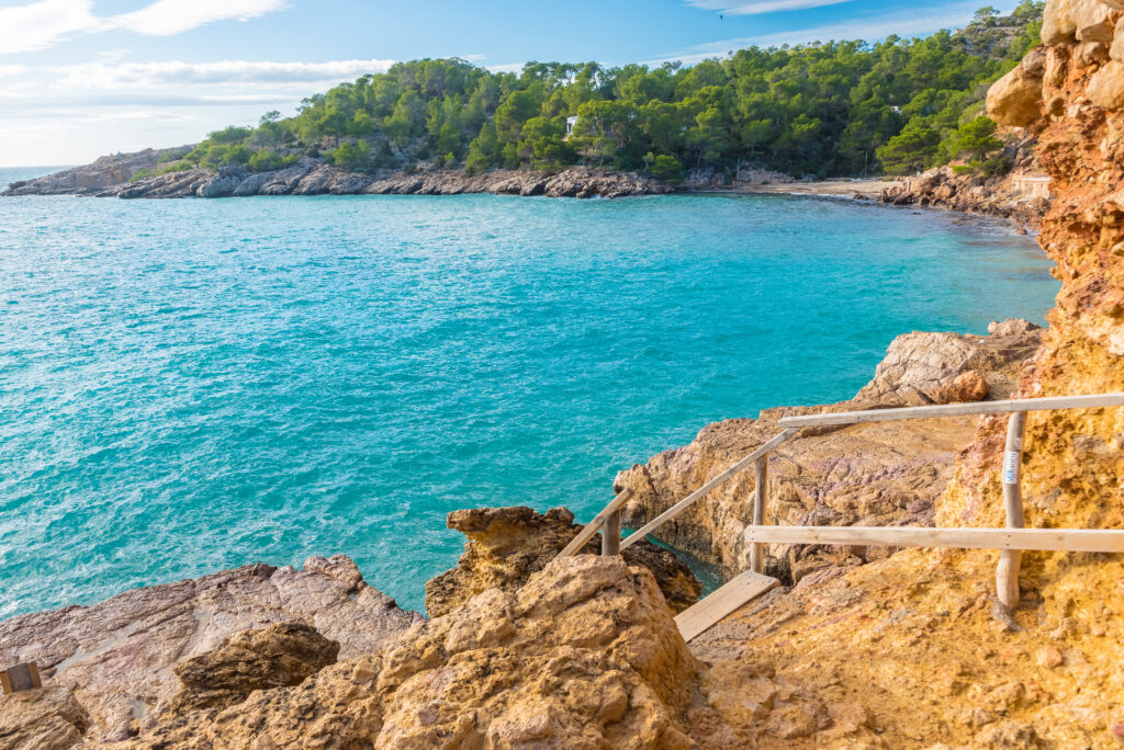 Discover the Spanish Costas with this coastal landscape featuring clear turquoise water, a rocky shore, wooden steps leading down to the beach, and a forested hill in the background under a partly cloudy sky.