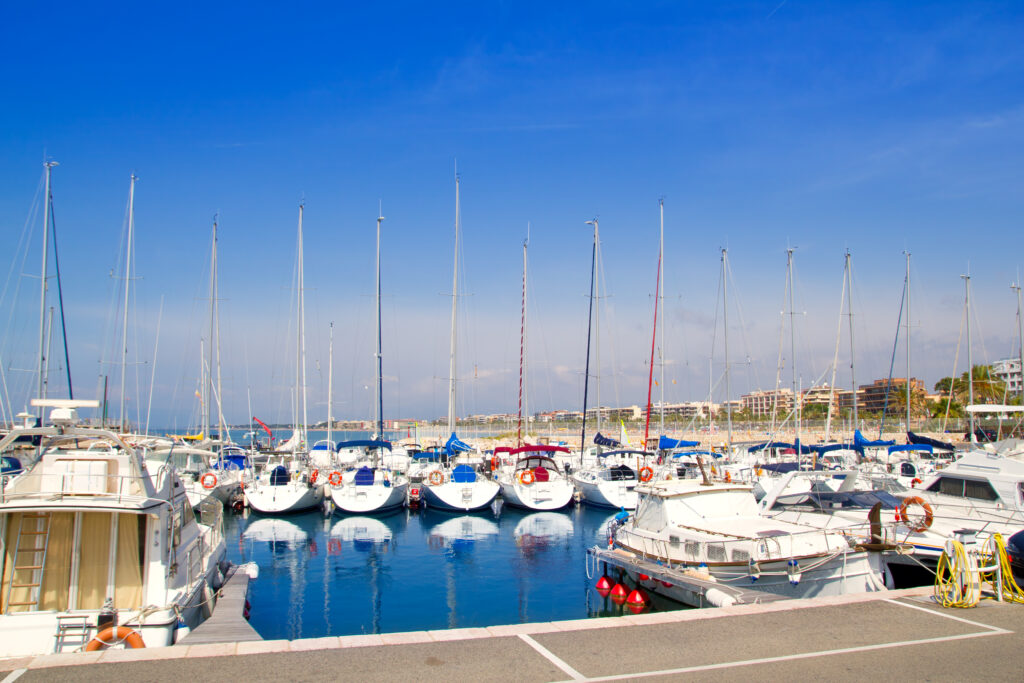 Discover the Spanish Costas as you admire a marina with multiple sailboats and yachts docked under a clear blue sky, apartment buildings, and palm trees framed beautifully in the background.