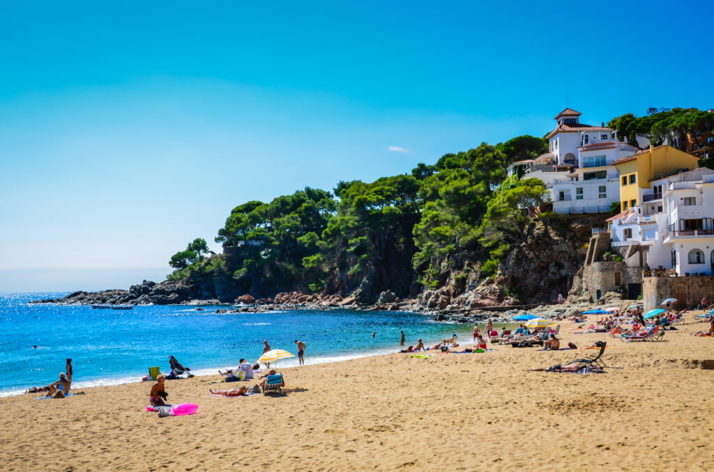 Discover the Spanish Costa Brava as you relax on a beach with a few people sunbathing and swimming. Nearby, houses and buildings perch atop a rocky cliffside with trees in the background, all under a clear blue sky.