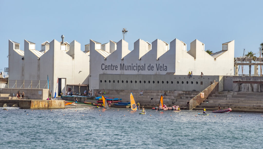 A vibrant scene at the Centre Municipal de Vela in Barcelona shows a bustling water sports facility with sailboats and kayaks along the waterfront on a clear day. People are actively engaged in various activities and enthusiastically discussing the upcoming America's Cup 2024 schedule. The nautical center serving as a hub for both recreational and competitive sailing enthusiasts.