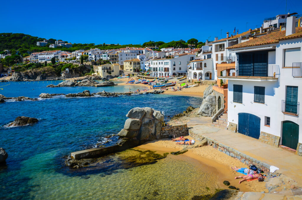 A coastal village on the enchanting Costa Brava features white houses and a sandy beach with sunbathers and boats. The clear blue sea and rocky outcrops are in the foreground.