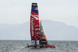 A red and black sailing catamaran with multiple sponsor logos, including Fly Emirates, sails on a calm sea with mountainous terrain in the background, as anticipation builds for America's Cup 2024 in Barcelona.