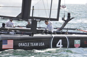 A man aboard an Oracle Team USA sailboat, labeled number 4, is adjusting equipment while racing on the water. The boat features U.S. flags and various sponsor logos, all set against the backdrop of excitement for America's Cup 2024.