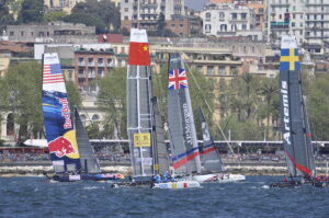 Four sailboats, representing China, the United Kingdom, Sweden, and Red Bull Racing, compete in a thrilling America's Cup Barcelona race on sparkling waters, with a vibrant cityscape and lush trees forming the picturesque backdrop.