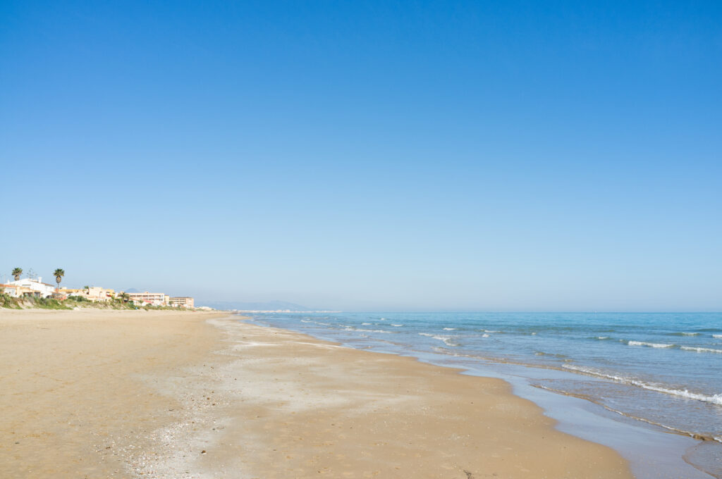 A wide sandy beach with gentle waves under a clear blue sky stretches along the Costa Blanca. Few buildings are visible in the distance.