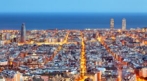 Aerial view of Barcelona city towards the sea, featuring notable landmarks such as Hotel Arts, Torre Agbar, and the area where the marina and the nautical school are located. This setting perfectly captures the environment for the upcoming America's Cup Barcelona.