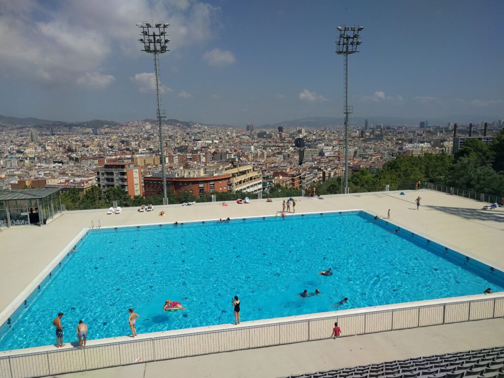 Outdoor swimming pool with people swimming and lounging around, situated in a spacious area with a view of the cityscape in the background. Tall stadium lights are positioned around the pool, and you can almost imagine the excitement of America's Cup Barcelona 2024 unfolding nearby.