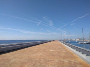 A long concrete pier with lampposts extends into a blue calm sea under a clear sky with crisscrossing contrails. Ships and boats, possibly preparing for the America’s Cup 2024, are seen in the distance.