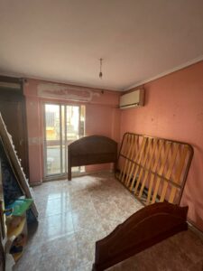 This Costa Dorada property for sale features an empty room with pink walls and a tiled floor. Disassembled bed frames are leaned against the walls, while a sliding glass door opens to a balcony. An air conditioning unit is mounted on the wall, adding modern comfort to this charming space.