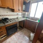 A charming kitchen with wooden cabinets, a gas stove, and granite countertops graces this Costa Dorada property for sale. The open cupboard offers easy access while cleaning supplies and a trash bag await tidying.