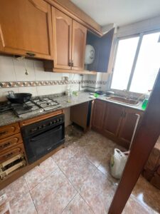 A charming kitchen with wooden cabinets, a gas stove, and granite countertops graces this Costa Dorada property for sale. The open cupboard offers easy access while cleaning supplies and a trash bag await tidying.