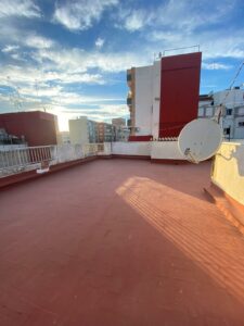 A flat rooftop with a red surface and a satellite dish overlooks the breathtaking cityscape of Costa Dorada, where buildings meet a partly cloudy sky at sunset—a perfect vista for those seeking property for sale.