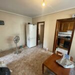 A small, sparsely furnished room in a Costa Dorada property for sale features a white fridge, fan, and table. The beige walls complement a door leading to the kitchen area with wooden cabinets. A clock hangs on one wall.