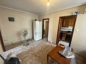 A small, sparsely furnished room in a Costa Dorada property for sale features a white fridge, fan, and table. The beige walls complement a door leading to the kitchen area with wooden cabinets. A clock hangs on one wall.