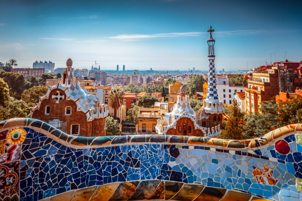 Colorful mosaic tiles on a curved bench overlook gingerbread-like buildings and a cityscape in Park Güell, Barcelona, under a clear blue sky—a perfect spot to ponder the ins and outs of Temporary rentals in Spain.