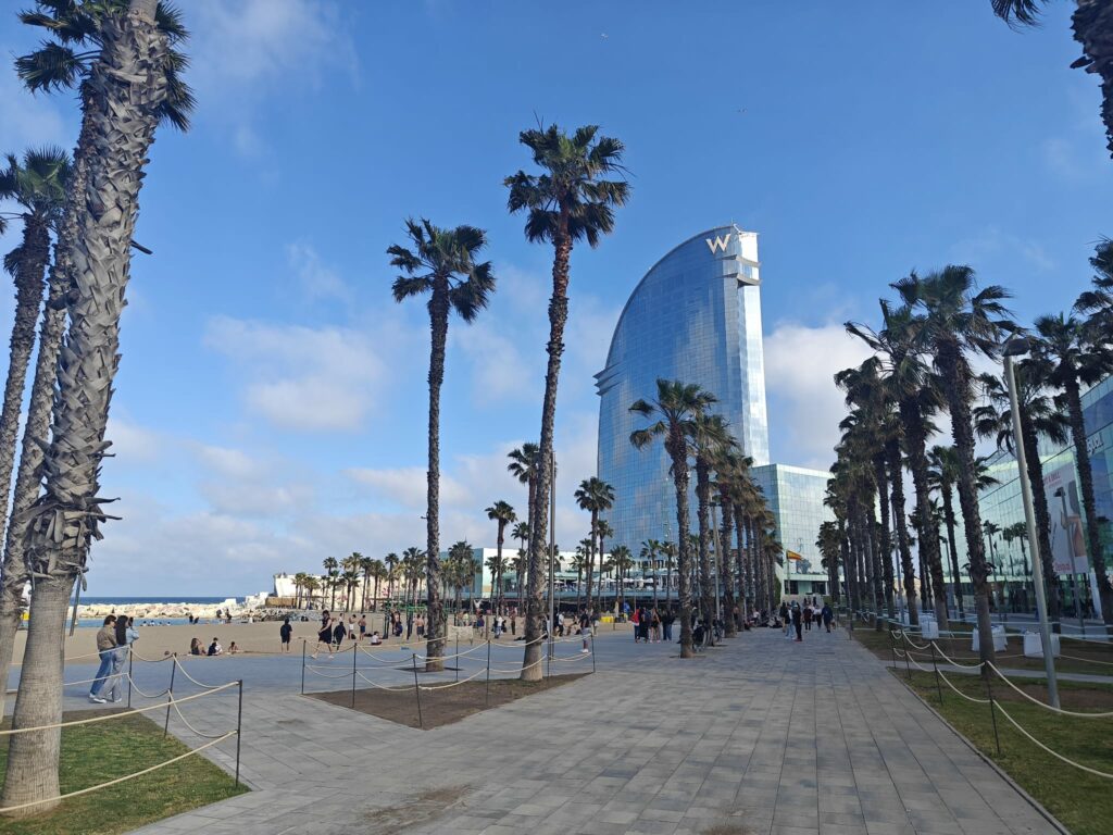 Palm trees line a walkway near a sandy beach bustling with people; a tall, sail-shaped glass building rises in the background—an iconic spot often featured in buying property in Spain guides and discussions on holiday rental Spain regulations.