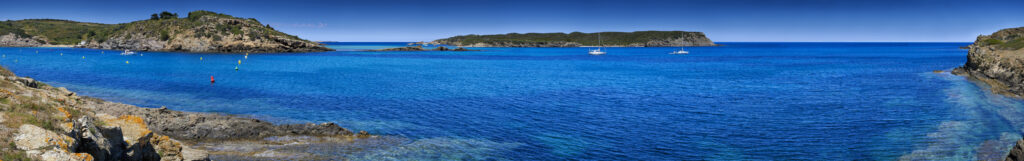 Panoramic view of a calm blue sea with a rocky coastline, small islands, and anchored sailboats under a clear sky—one of the best things to do in Balearic Islands for nature lovers and sailing enthusiasts.