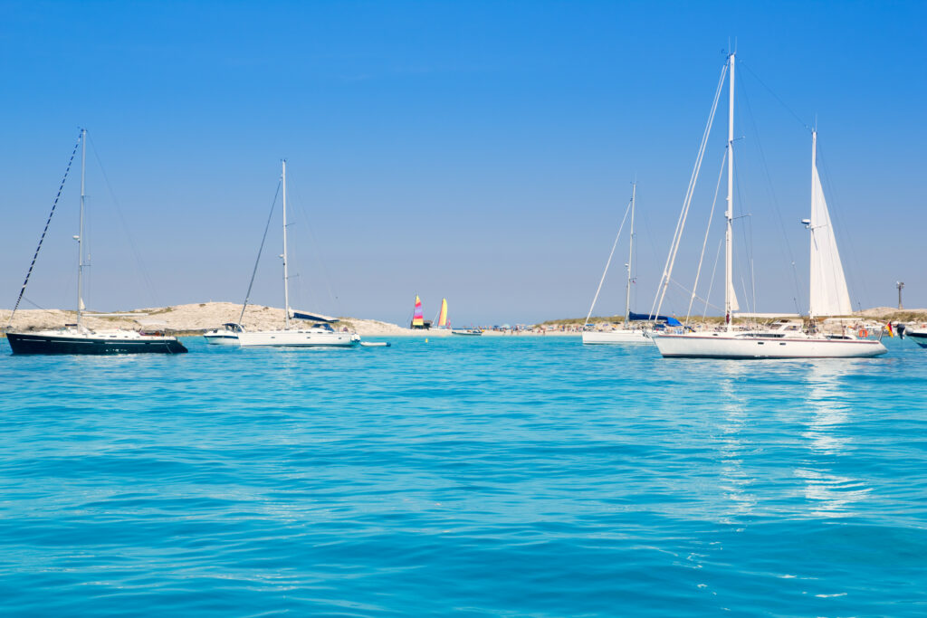 Several sailboats float on calm, clear blue water near a sandy shoreline under a bright blue sky, evoking the tranquil beauty often found in the Balearic Islands.