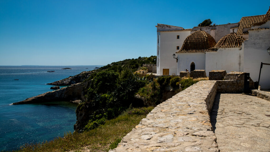 A stone path winds along a coastal cliff beside a white building with domed roofs, overlooking the blue sea—a scene straight from the Balearic Islands map and one of the best things to do in the Balearic Islands.